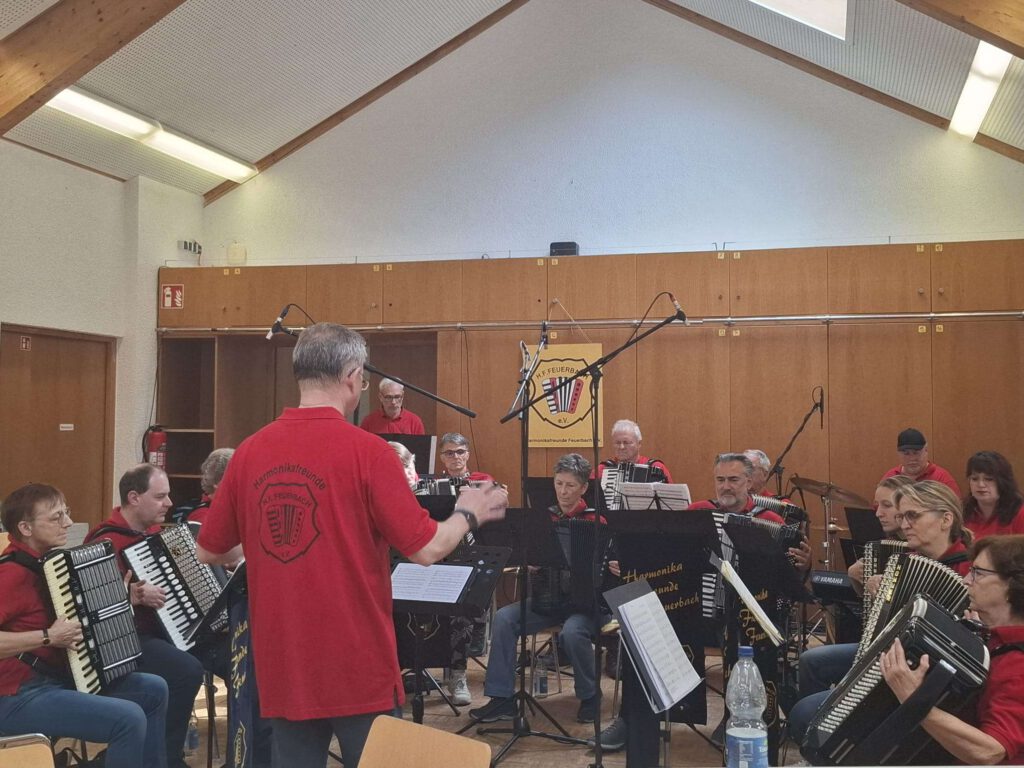 Auf dem Bild ist das 1. Orchester der Harmonikafreunde Feuerbach beim Auftritt im großen Saal des Waldheim Lindentals zu sehen. Insgesamt 15 Spieler und Spielerinnen unterschiedlichen Alters sitzen in einem Halbkreis mit Akkordeons auf dem Schoß. Vor ihnen stehen Notenständer, an denen dunkelblaue Banner mit der Aufschrift "Harmonikafreunde Feuerbach" und dem HFF Logo (ein Akkordeon in einem Wappen) angebracht sind. Alle tragen rote Poloshirts. Vor dem Orchester steht Vitali Neiffert mit dem Rücken zum Publikum. Auch er trägt ein rotes Poloshirt. Auf seinem Rücken ist das Logo des HFF zu sehen. Er hat beide Hände zum Dirigieren in der Luft und schaut auf die Noten vor sich. Im Hintergrund ist eine Schrankwand zu sehen, an der das Wappen des HFF angebracht ist.