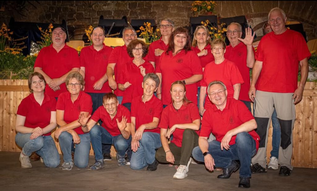 Ein Gruppenfoto des ersten Orchesters. Abgebildet sind 16 Personen unterschiedlichen Alters, alle mit roten HFF T-Shirts. Sie sind in 3 Reihen aufgestellt, die vorderste Reihe kniet auf dem Boden. Alle lachen in die Kamera. Im Hintergrund ist eine aufgebaute Bühne mit Blumendekoration zu sehen.