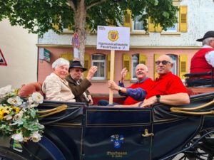 Auf dem Bild ist eine schwarze Kutsche zu sehen. In ihr sitzen vier Personen, drei Männer und eine Frau. Einer der Männer, Reiner Hofsäß, der 1. Vorsitzende des HFF, hält ein Schild mit dem HFF Logo hoch.
