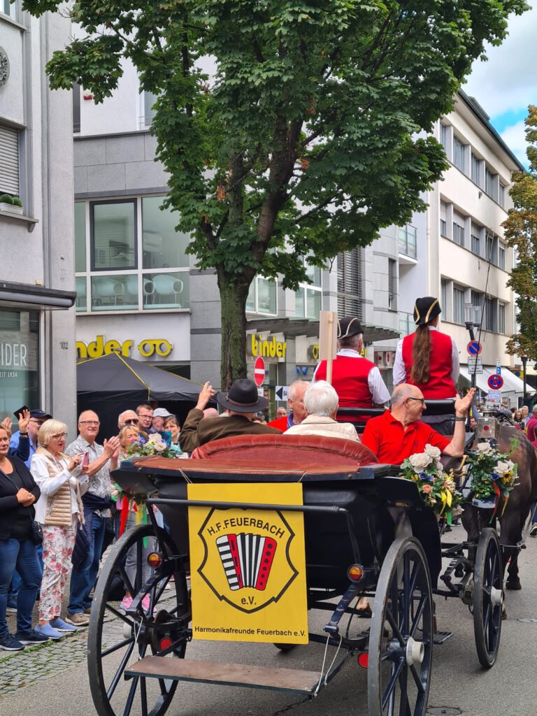 Eine schwarze Kutsche ist von hinten zu sehen. Sie ist mit Blumen geschmückt. Prominent hängt an ihr das Logo der Harmonikafreunde Feuerbach, ein rotes Akkordeon auf gelbem Grund. In der Kutsche sitzen vier Mitglieder des HFF und winken. Am Rand der Straße sind jubelnde Zuschauer zu sehen.