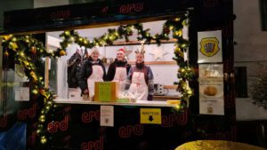 Ein Anhänger mit großem Ausgabefenster ist mit einer Girlande aus Tannenzweigen, Lichtern, und Weihnachtsdekorationen geschmückt. An dem Wagen, der mit dem Logo des ERD bedruckt ist, hängt das Logo der Harmonikafreunde Feuerbach, sowie verschiedene Zettel, die Waffeln, Kekse, und Fleischkäse anpreisen. In dem Wagen hängen ebenfalls festliche Dekorationen. Drei Mitglieder des HFF stehen in dem Wagen mit weißen Schürzen, auf denen in rot das Logo des HFF abgebildet ist. Eine trägt ein Geweih, eine andere eine Weihnachtsmütze. Sie lächeln in die Kamera.