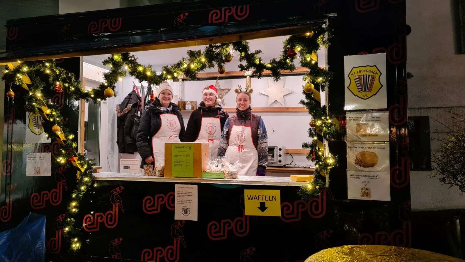 Ein Anhänger mit großem Ausgabefenster ist mit einer Girlande aus Tannenzweigen, Lichtern, und Weihnachtsdekorationen geschmückt. An dem Wagen, der mit dem Logo des ERD bedruckt ist, hängt das Logo der Harmonikafreunde Feuerbach, sowie verschiedene Zettel, die Waffeln, Kekse, und Fleischkäse anpreisen. In dem Wagen hängen ebenfalls festliche Dekorationen. Drei Mitglieder des HFF stehen in dem Wagen mit weißen Schürzen, auf denen in rot das Logo des HFF abgebildet ist. Eine trägt ein Geweih, eine andere eine Weihnachtsmütze. Sie lächeln in die Kamera.