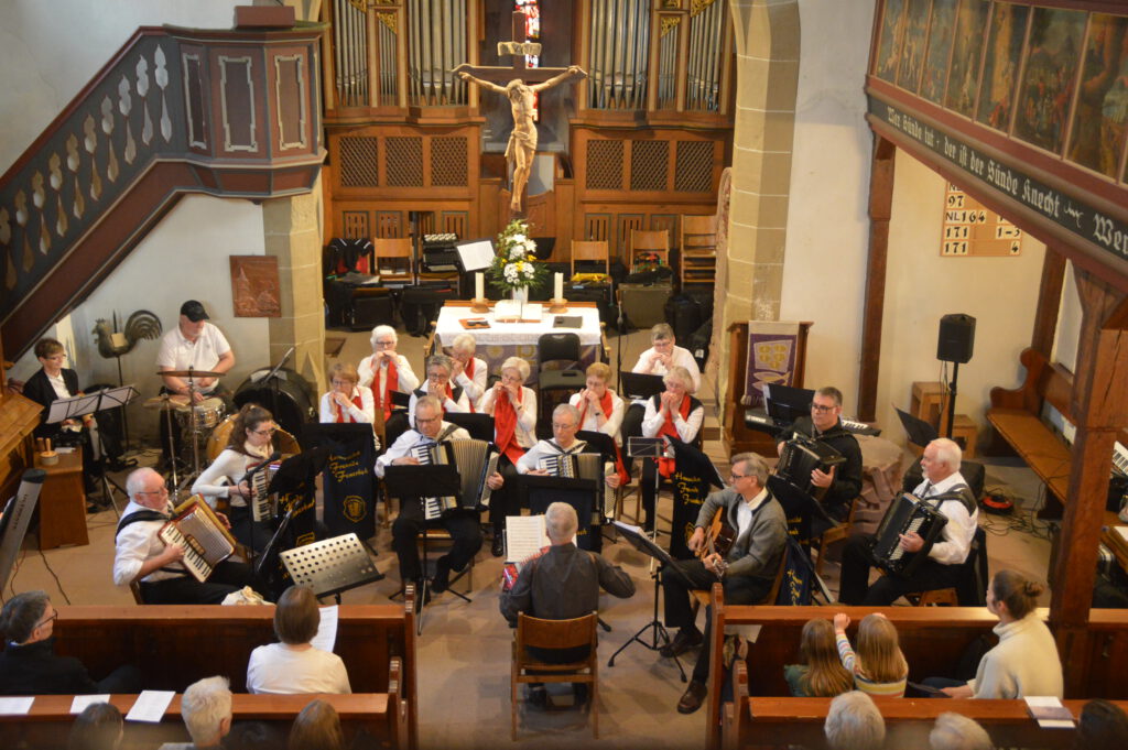 Die Volksmusikgruppe, gekleidet in weiß-schwarz, sitzt mit Instrumenten vor dem Altar der Oswaldkirche Weilimdorf. Sie werden verstärkt durch Mitglieder des Cannstatter Mundharmonikaorchesters mit roten Schals in der 2. und 3. Reihe. Vorne sitzt Walter Krebs mit dem Rücken zum Publikum und einer kleinen Handharmonika auf dem Schoß.