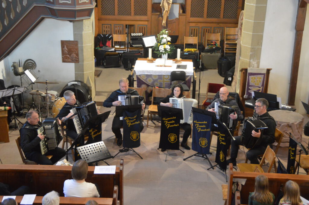 Das Ensemble der Harmonikafreunde, bestehend aus 6 Spielenden, sitzt mit Instrumenten vor dem Altar der Oswaldkirche. Sie sind in ihr Spiel vertieft.