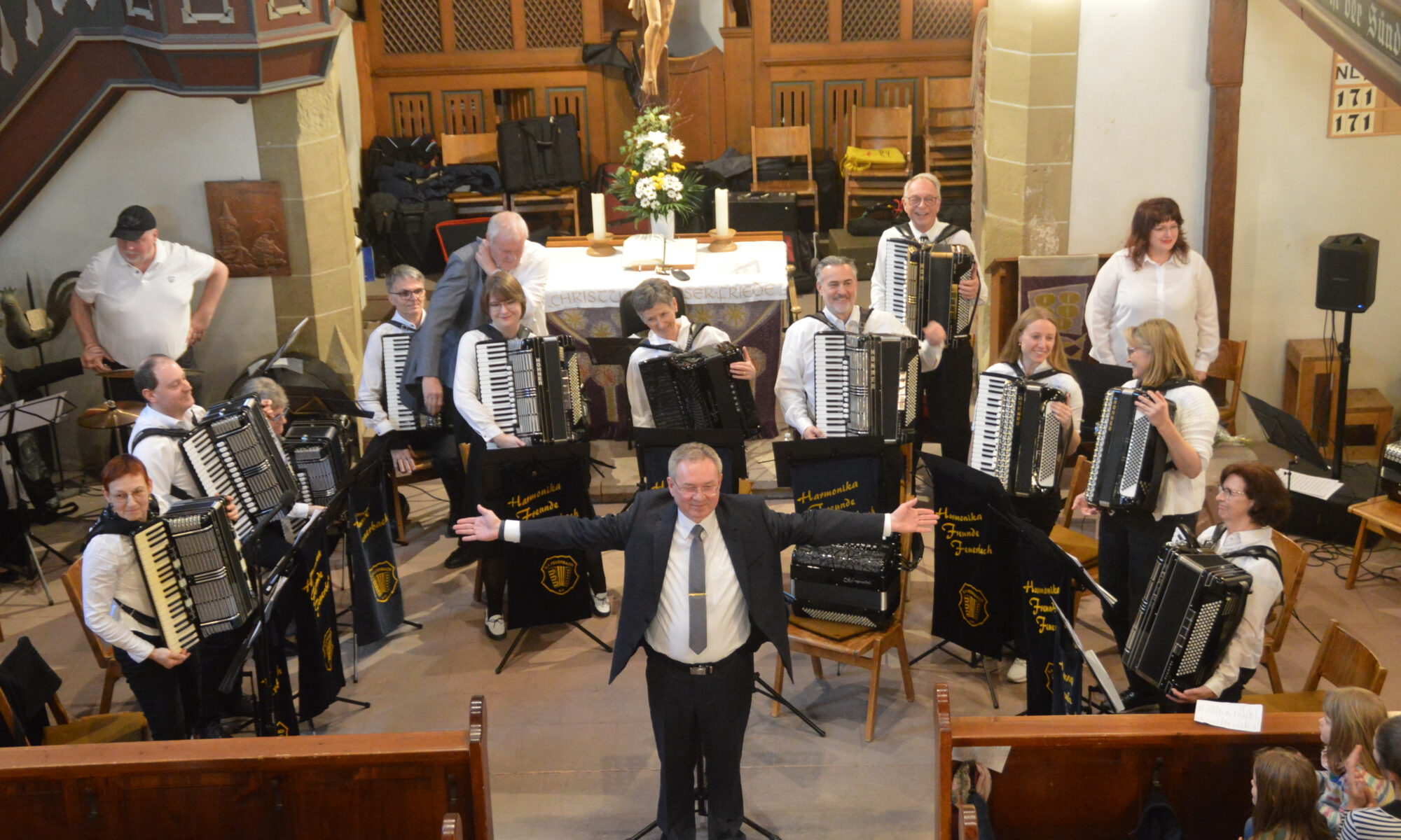 Das 1. Orchester der Harmonikafreunde Feuerbach steht vor dem Altar der Oswaldkirche und nimmt den Applaus des Publikums entgegen. Vorne steht ihr Dirigent Vitali Neiffert in Anzug und Krawatte, die Arme ausgebreitet, ein Lächeln im Gesicht.