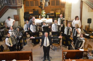 Das 1. Orchester der Harmonikafreunde Feuerbach steht vor dem Altar der Oswaldkirche und nimmt den Applaus des Publikums entgegen. Vorne steht ihr Dirigent Vitali Neiffert in Anzug und Krawatte, die Arme ausgebreitet, ein Lächeln im Gesicht.