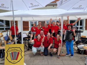 Das 1. Orchester der Harmonikafreunde Feuerbach winkt fröhlich in die Kamera. Alle tragen rote HFF T-Shirts. Im Hintergrund ist das Waldheim Lindental zu sehen.